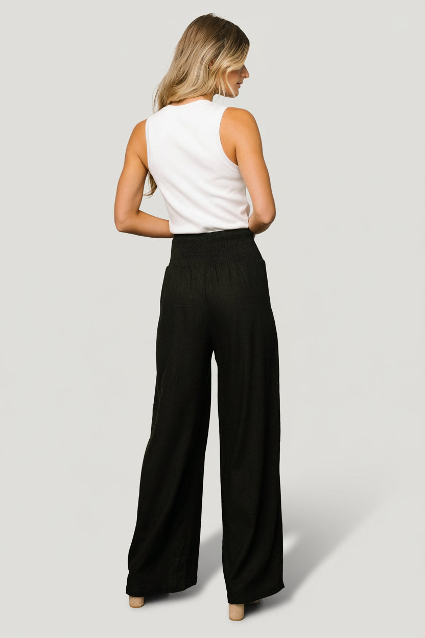 Full-length styling image of Parvati black smocked waist pants, highlighting side pockets and relaxed wide-leg cut.