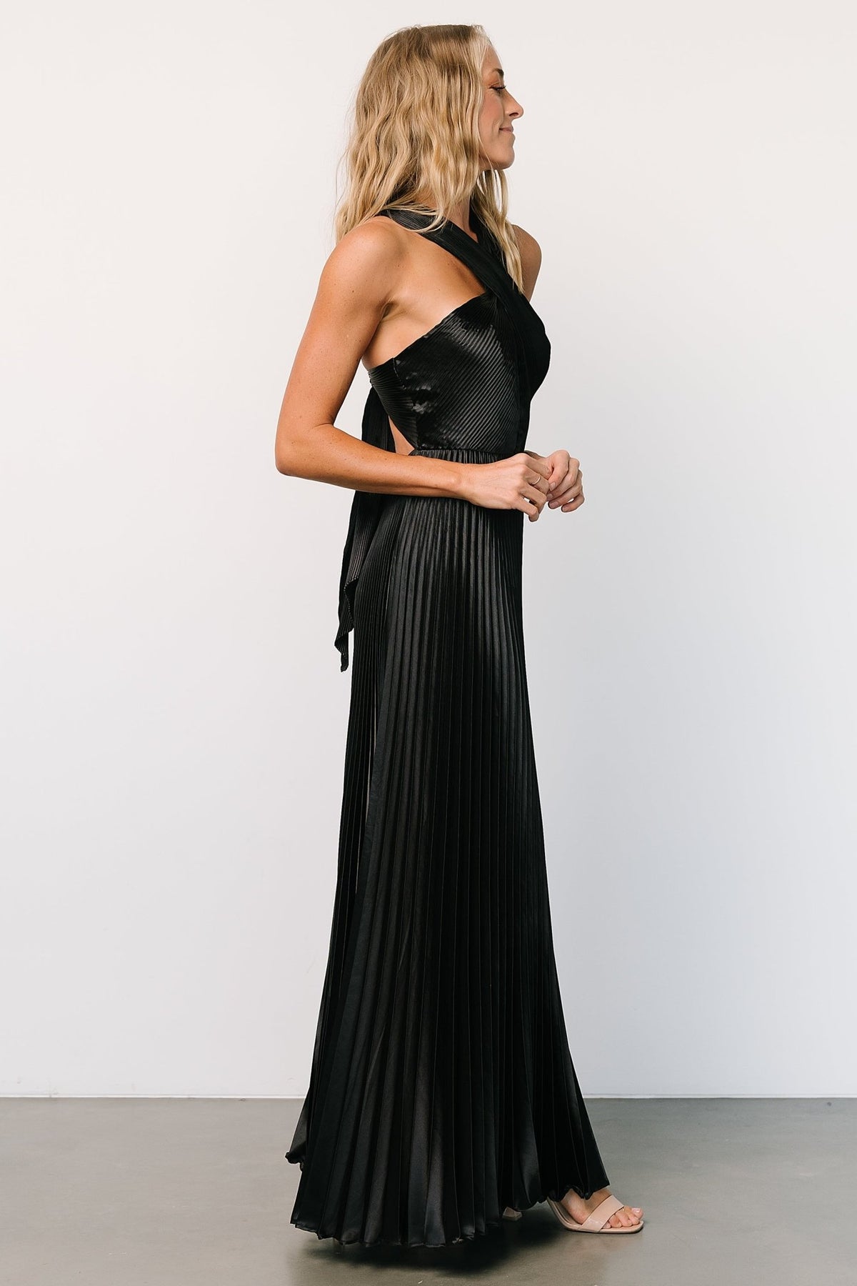 Studio shot of Aakaa Sandra pleated maxi dress in black with soft pleats and fluid drape, styled with minimal accessories.