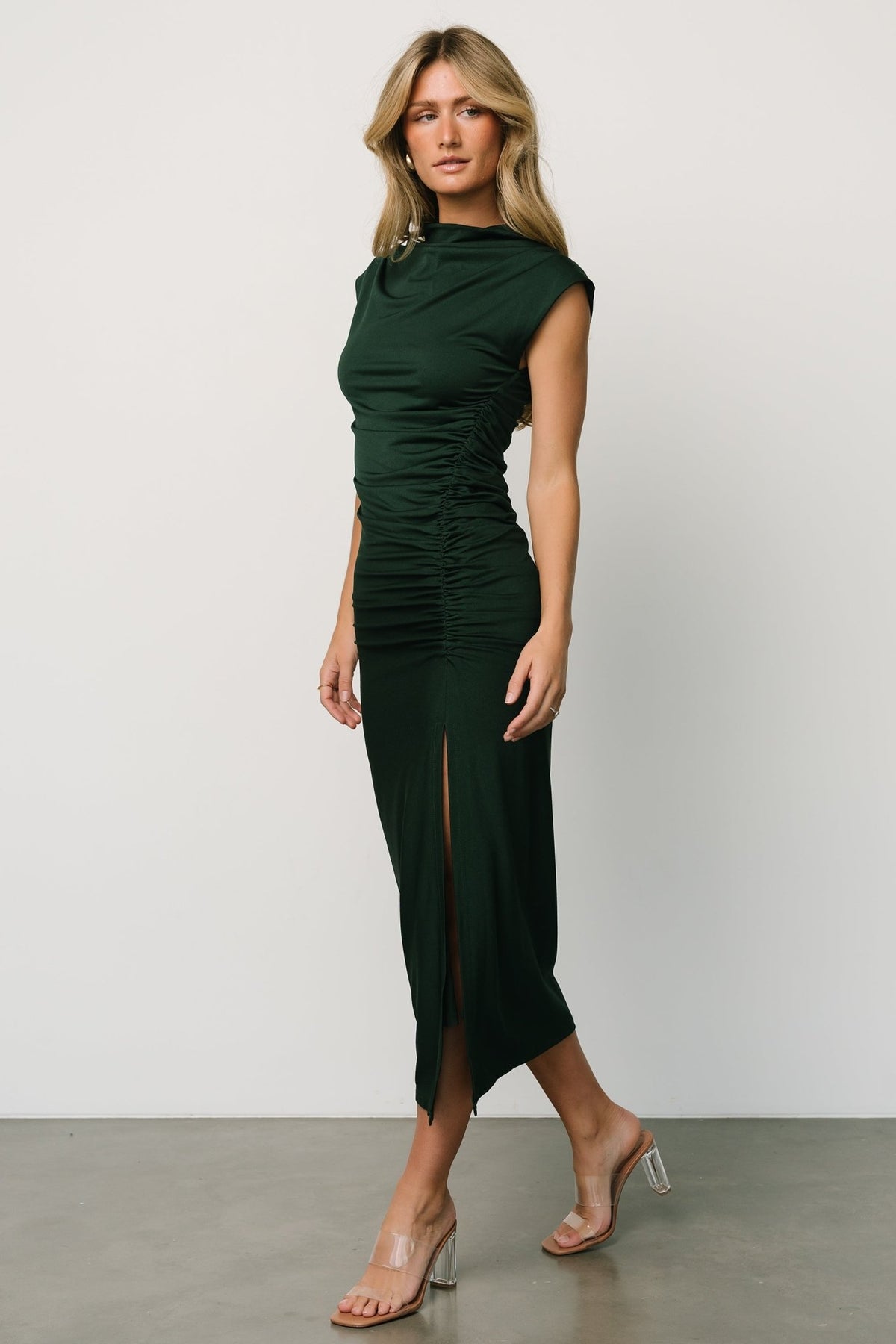 Model walking in Sasha Ruched Maxi Dress in deep green, showing movement, cowl neckline, short cap sleeves and left leg slit.