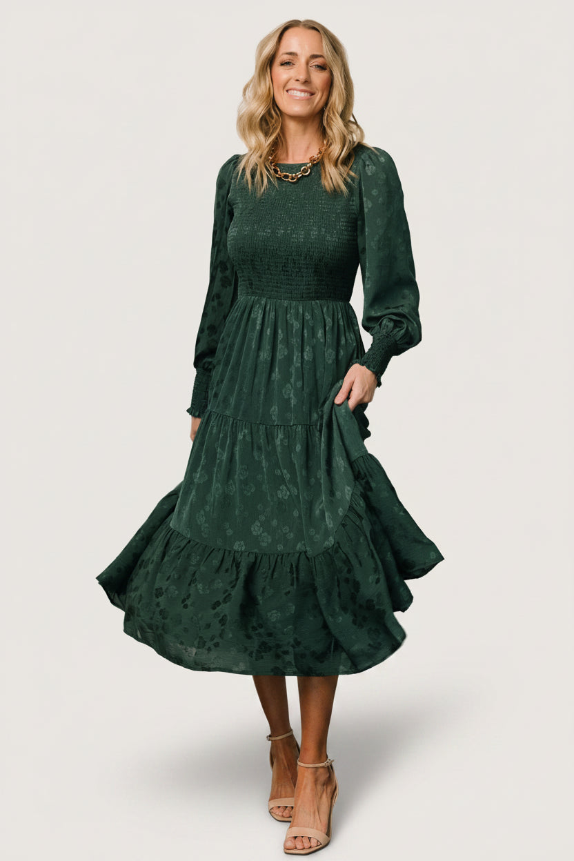 Front view of Stockholm Smocked Dress in dark hunter green with subtle shiny floral pattern, smocked bodice and tiered midi skirt, on model.