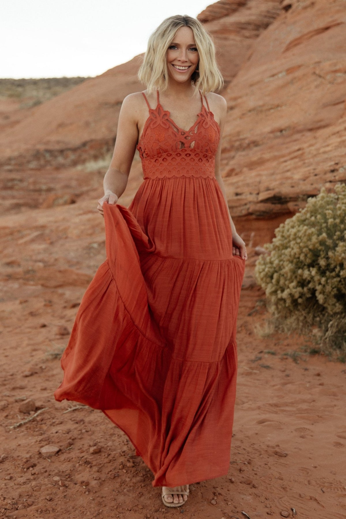 Rust lace-bodice maxi dress on model showing adjustable double criss-cross straps and tiered skirt in motion, fully lined.
