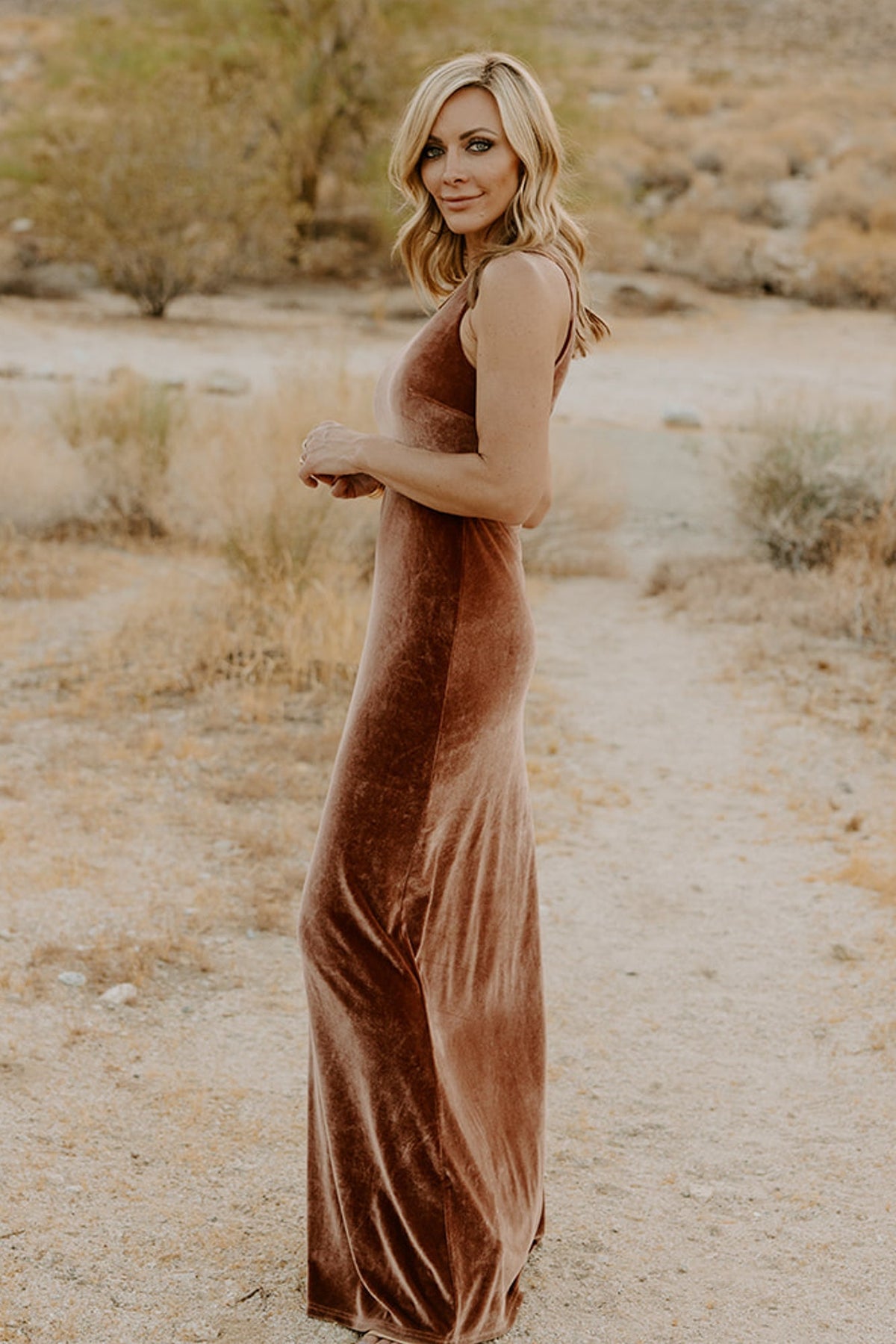 Angled model pose in bronze velvet maxi highlighting fitted waist, asymmetrical neckline, and graceful skirt movement with side slit visible.